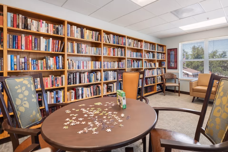 A cozy library room with wooden bookshelves filled with books along the wall. In the foreground, there is a round wooden table with a partially completed jigsaw puzzle and a puzzle box on it. Surrounding the table are several chairs with patterned cushions. The room has a large window letting in natural light and additional seating including an armchair and a wooden chair near the window.