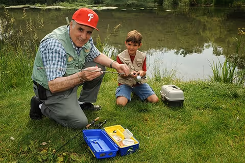 An older man wearing a red baseball cap and a fishing vest is kneeling on the grass next to a young boy who is also kneeling. They are by the edge of a pond, holding a fishing rod together. A blue tackle box and a small container are on the grass nearby.
