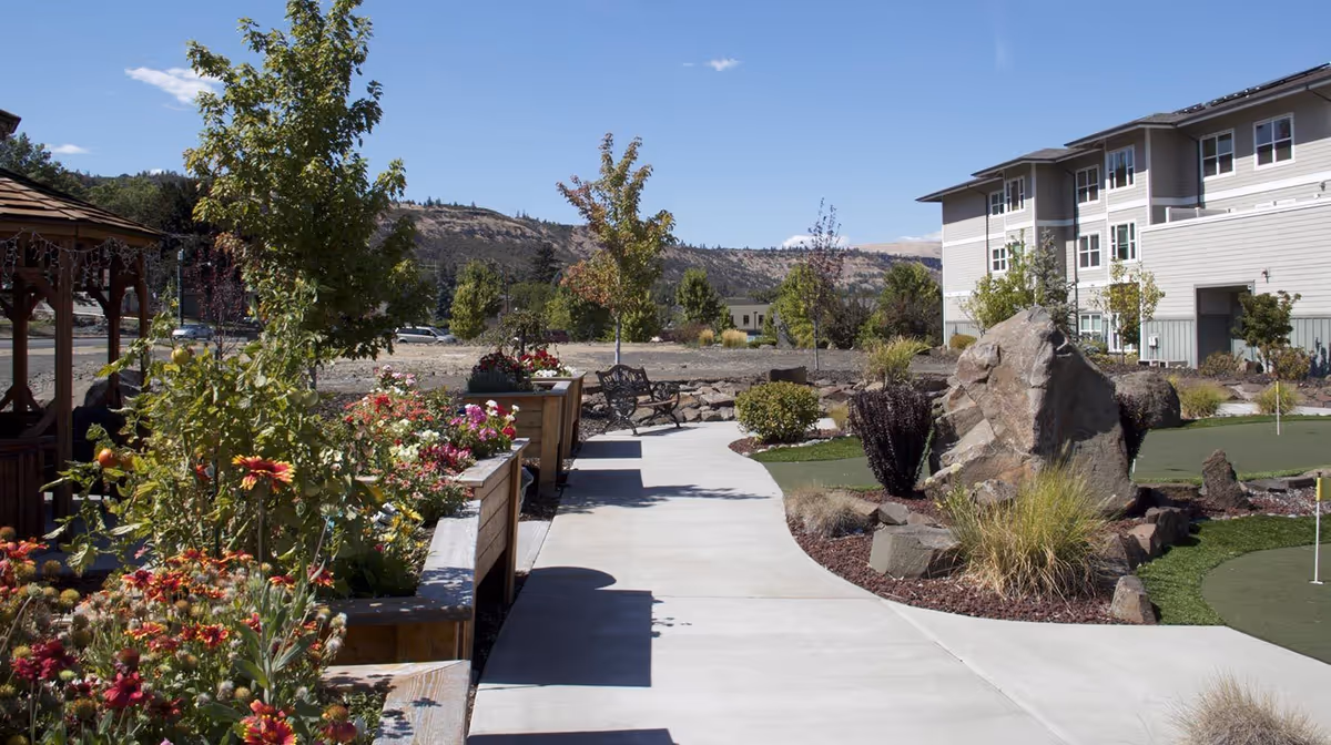 Sunlit outdoor courtyard at a senior living community with a paved walkway, flower beds, benches, and a building in the background.