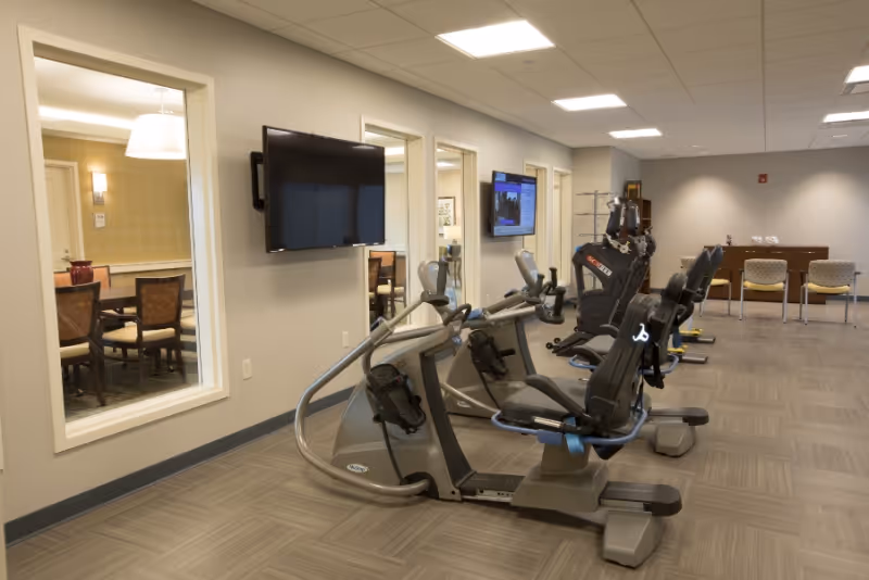 A fitness room in a senior living facility with several exercise bikes lined up facing a wall-mounted TV. The room has large windows looking into an adjacent dining area with a table and chairs. The floor is covered with gray tiles, and the walls are painted light gray. There are also chairs and a table against the far wall.
