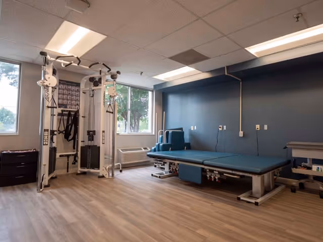A rehabilitation room with exercise equipment including a multi-functional weight machine and a padded treatment table. The room has large windows letting in natural light, wood-style flooring, and a blue accent wall.