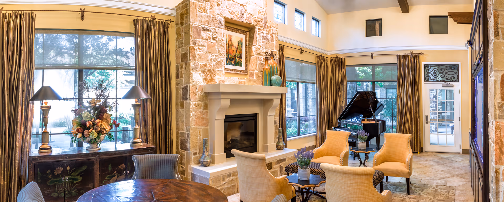 A spacious and well-lit living room featuring large windows with brown curtains, a stone fireplace with decorative vases and a framed painting above it, a round wooden table with chairs, a grand piano near the windows, and comfortable beige armchairs arranged around a small table with potted plants.