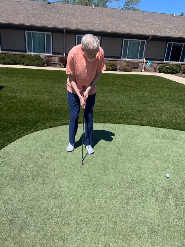 An elderly man wearing a pink striped shirt and blue pants is playing mini golf on a putting green outside a single-story building with windows and bushes in the background.