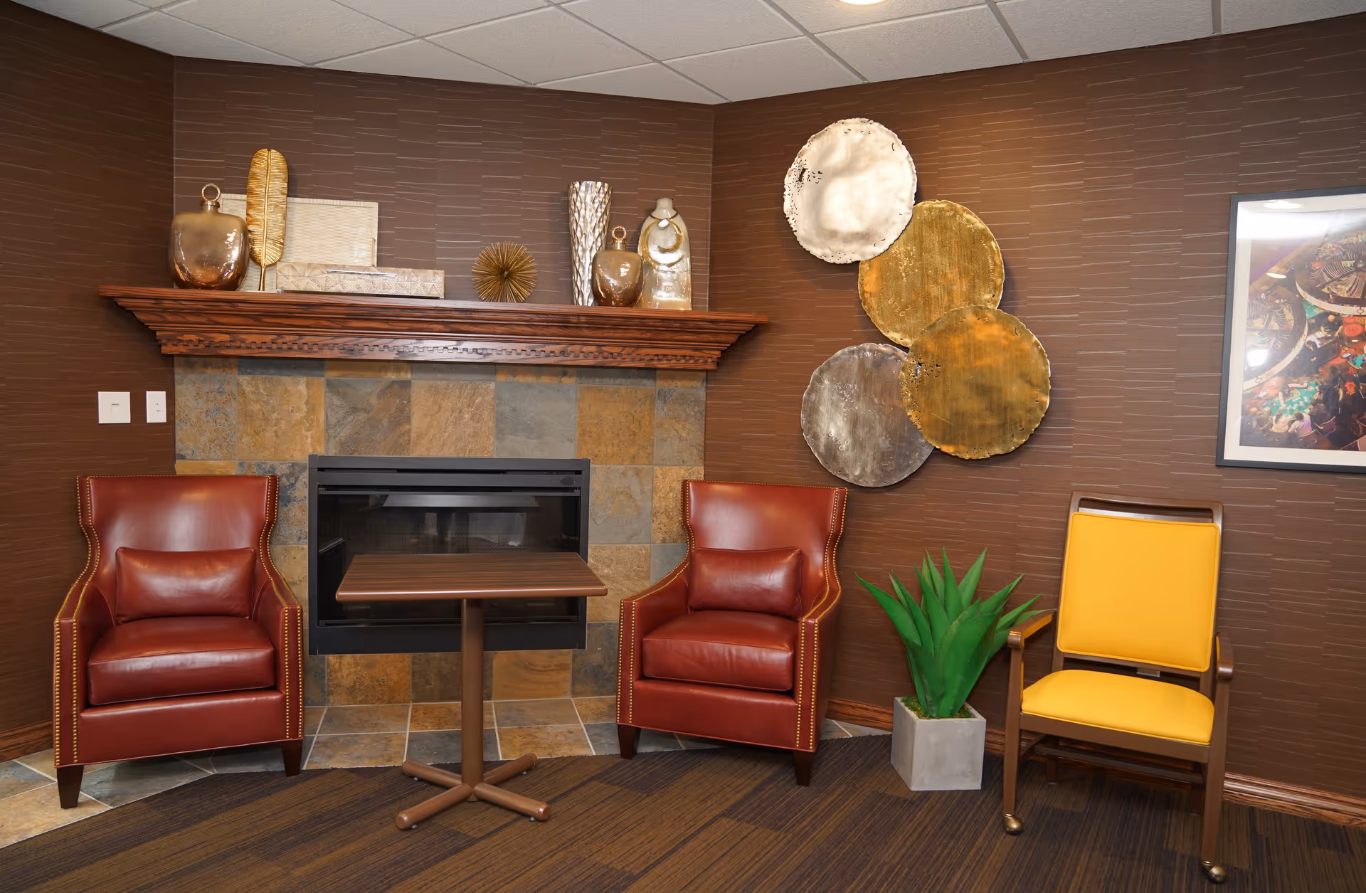 A cozy corner in a retirement community featuring two red leather armchairs with nailhead trim, a small wooden table between them, and a yellow cushioned chair on wheels. The background includes a fireplace with a tiled surround and a wooden mantel decorated with vases and decorative objects. The walls are brown with textured wallpaper, adorned with a set of four metallic circular wall art pieces and a framed colorful picture. A green potted plant sits on the floor next to the yellow chair.