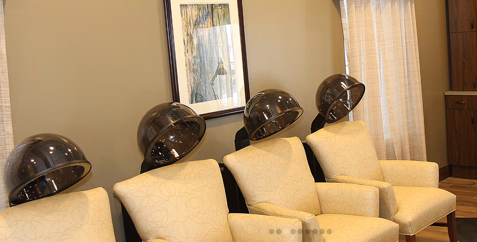Interior view of a salon area with four beige cushioned chairs, each positioned under a black hair dryer hood. A framed artwork hangs on the wall behind the chairs, and light-colored curtains cover the window to the right.