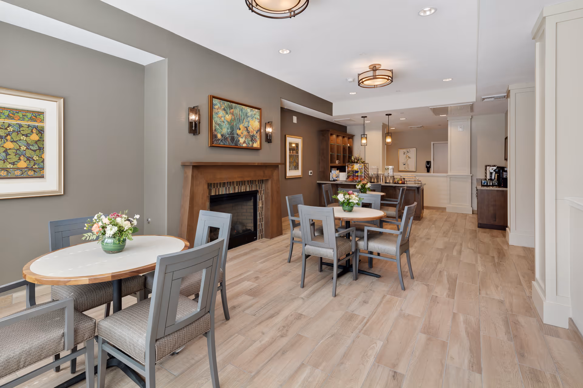 A bright and spacious dining area with light wood flooring, gray walls, and modern ceiling lights. The room features two round tables with gray chairs, each table decorated with a small flower arrangement. There is a wooden fireplace with artwork above it on the left wall, and a counter area with pendant lights and refreshments in the background.