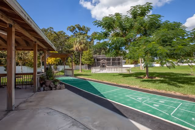 Outdoor area at The Goldton at St. Petersburg featuring a covered patio with wooden posts, a shuffleboard court with green playing surface, lush green grass, several trees including a palm tree, and a fenced enclosure in the background under a partly cloudy sky.