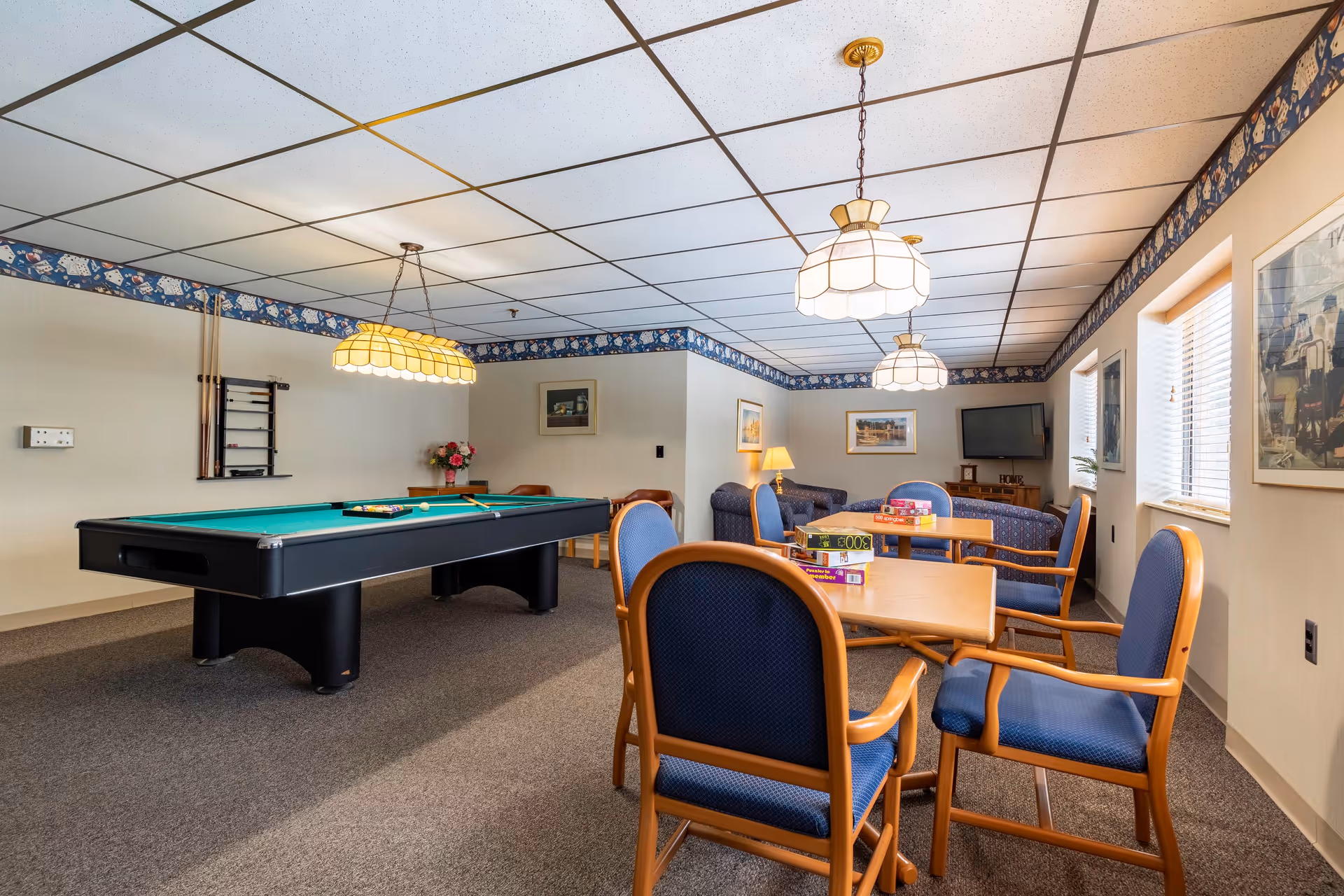 A recreational room with a pool table, several tables with blue cushioned chairs, board games on one table, a couch, a TV mounted on the wall, and framed pictures. The room has a drop ceiling with hanging lights and windows letting in natural light.