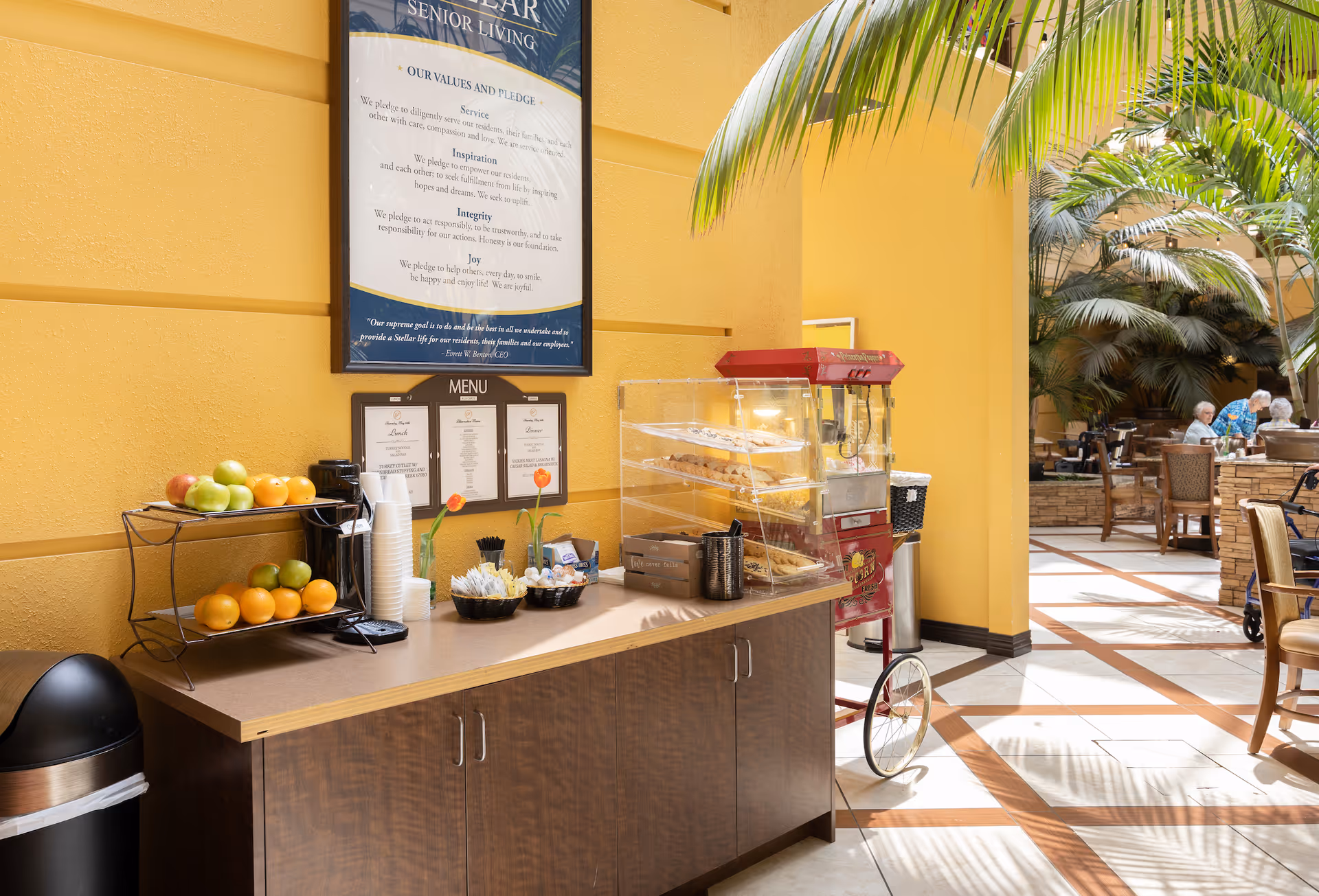 A bright senior living dining area with a snack station holding fruit, cups, and a popcorn machine, and seating with residents in the background.