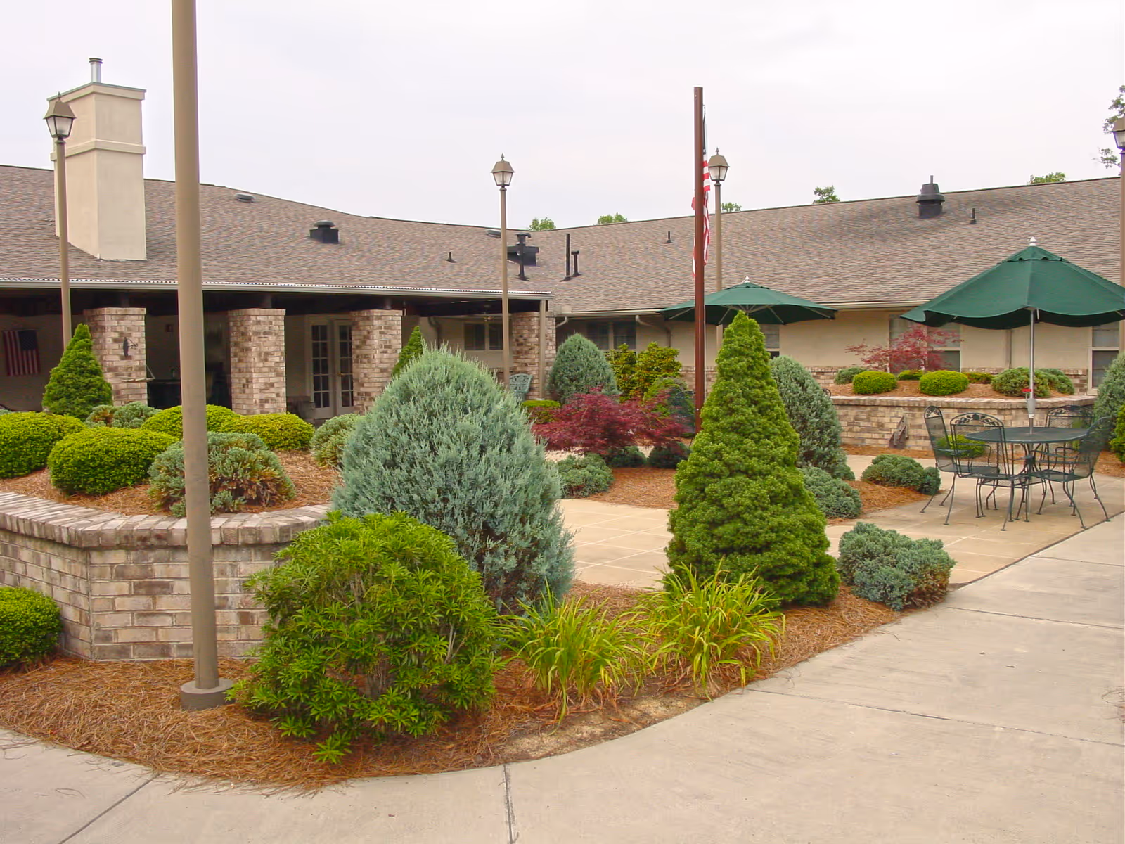 Outdoor courtyard area of a senior living facility with neatly trimmed bushes, small trees, and landscaped garden beds. There are green patio umbrellas over metal tables and chairs on a paved surface. The building with a shingled roof and brick columns surrounds the courtyard.