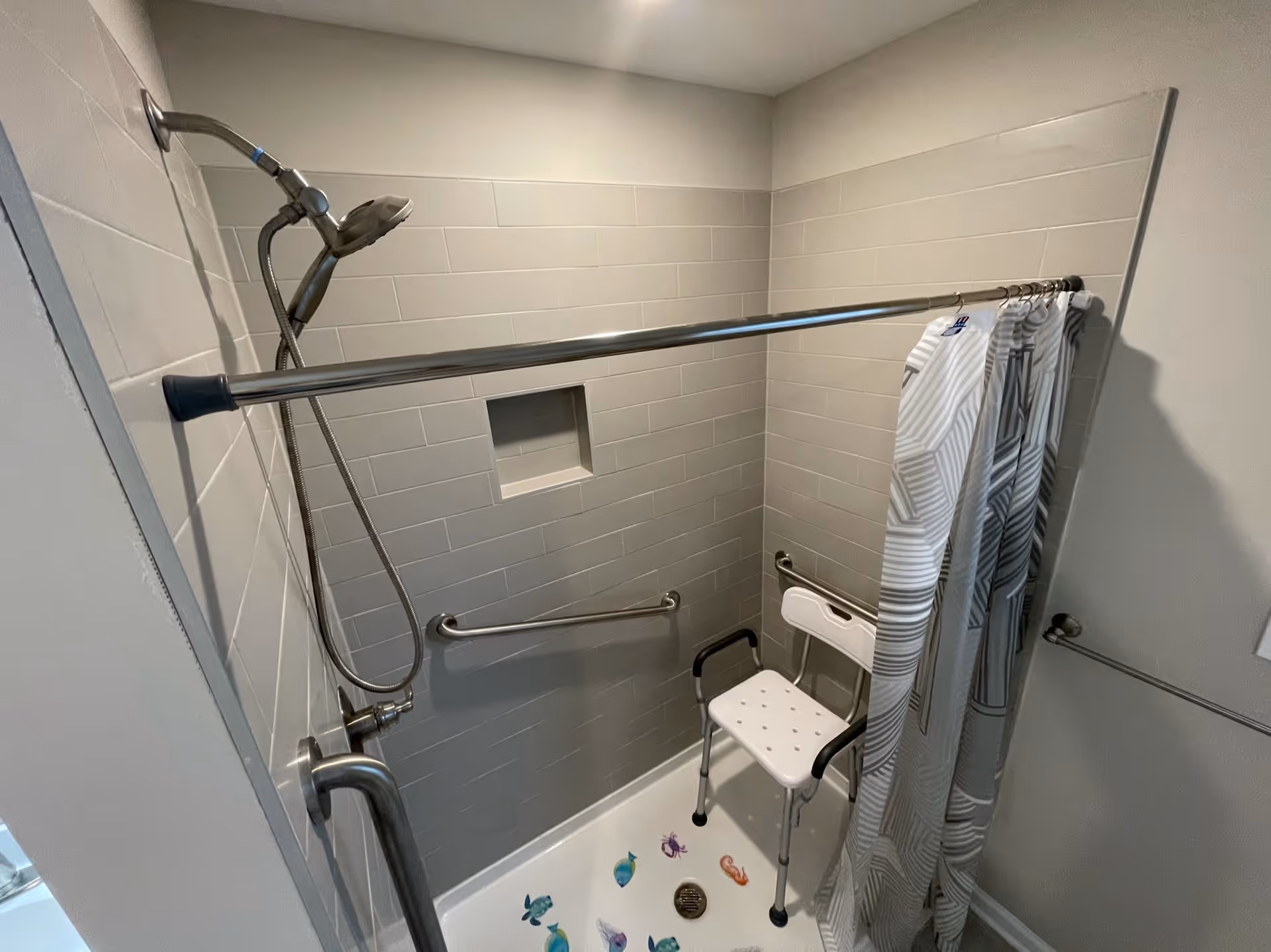 Tiled accessible shower with a handheld showerhead, grab bars, a white shower chair and a patterned curtain.