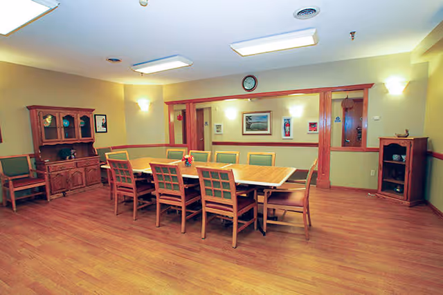 A dining room with a long wooden table surrounded by ten wooden chairs with green cushions. The room has wooden flooring, light yellow walls with a wooden chair rail, and several framed pictures on the walls. There is a wooden cabinet with glass doors on the left side and a smaller wooden cabinet on the right side. The ceiling has recessed lighting and a clock is mounted above the doorway.