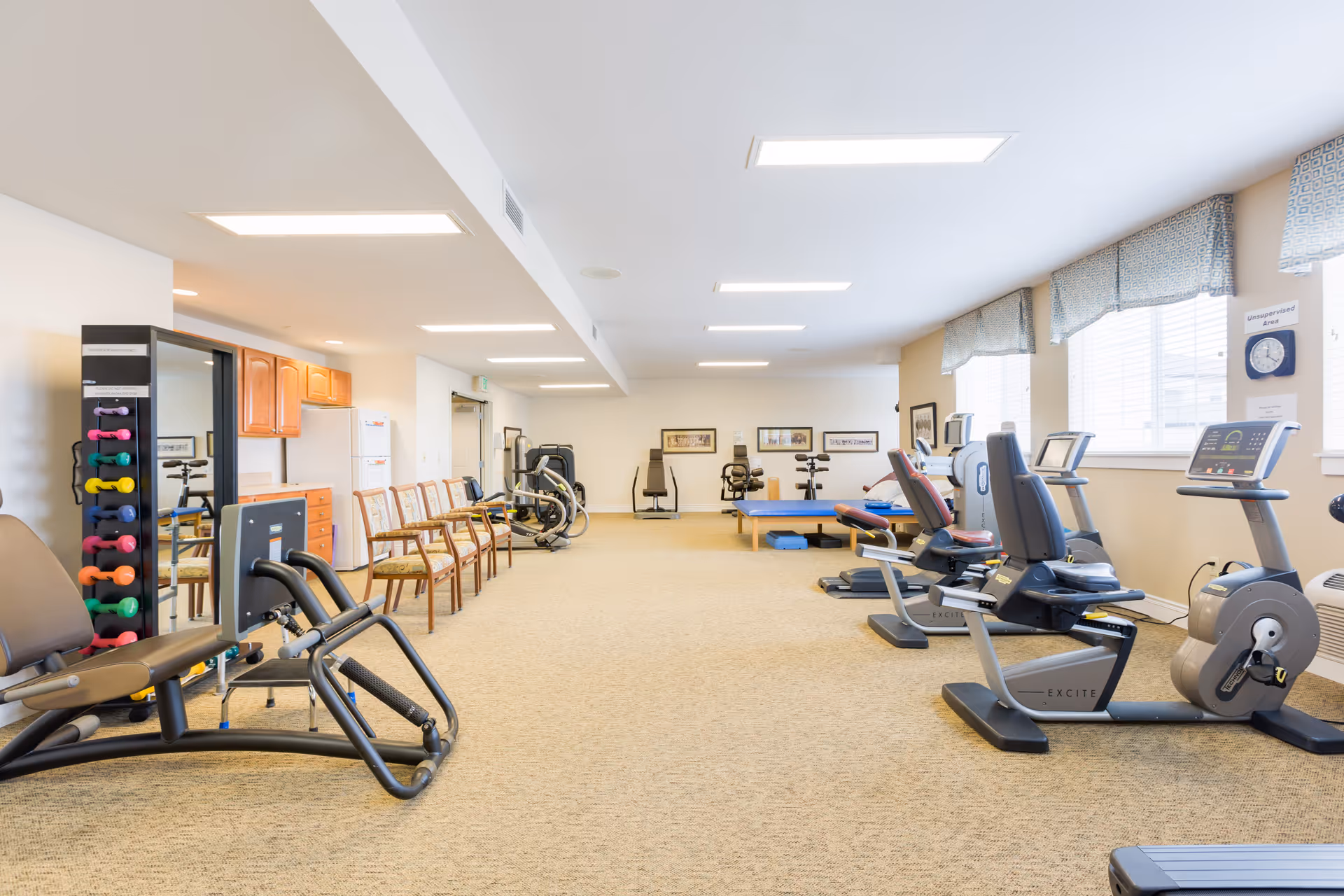 A spacious senior living fitness room with various exercise equipment including stationary bikes, weight machines, and a rack of colorful dumbbells. There are chairs lined up against the wall and large windows with patterned valances letting in natural light.