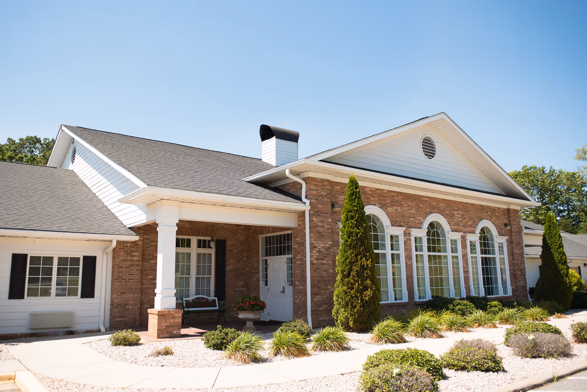 Exterior view of a single-story assisted living facility building with a brick facade, white trim, large arched windows, a covered entrance with white columns, and landscaped bushes and plants along the walkway.
