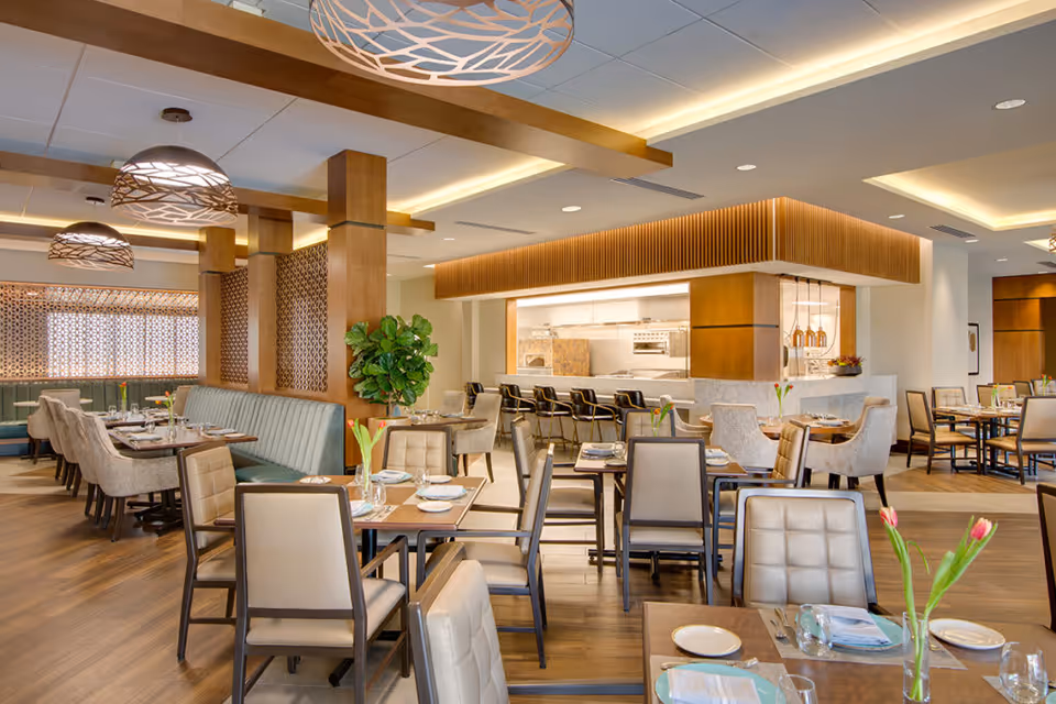 A spacious, modern dining room with multiple tables and chairs arranged neatly. The room features wooden flooring, decorative hanging light fixtures, and a large open kitchen area with bar stools. The tables are set with plates, glasses, and napkins, and there are small vases with flowers on each table.