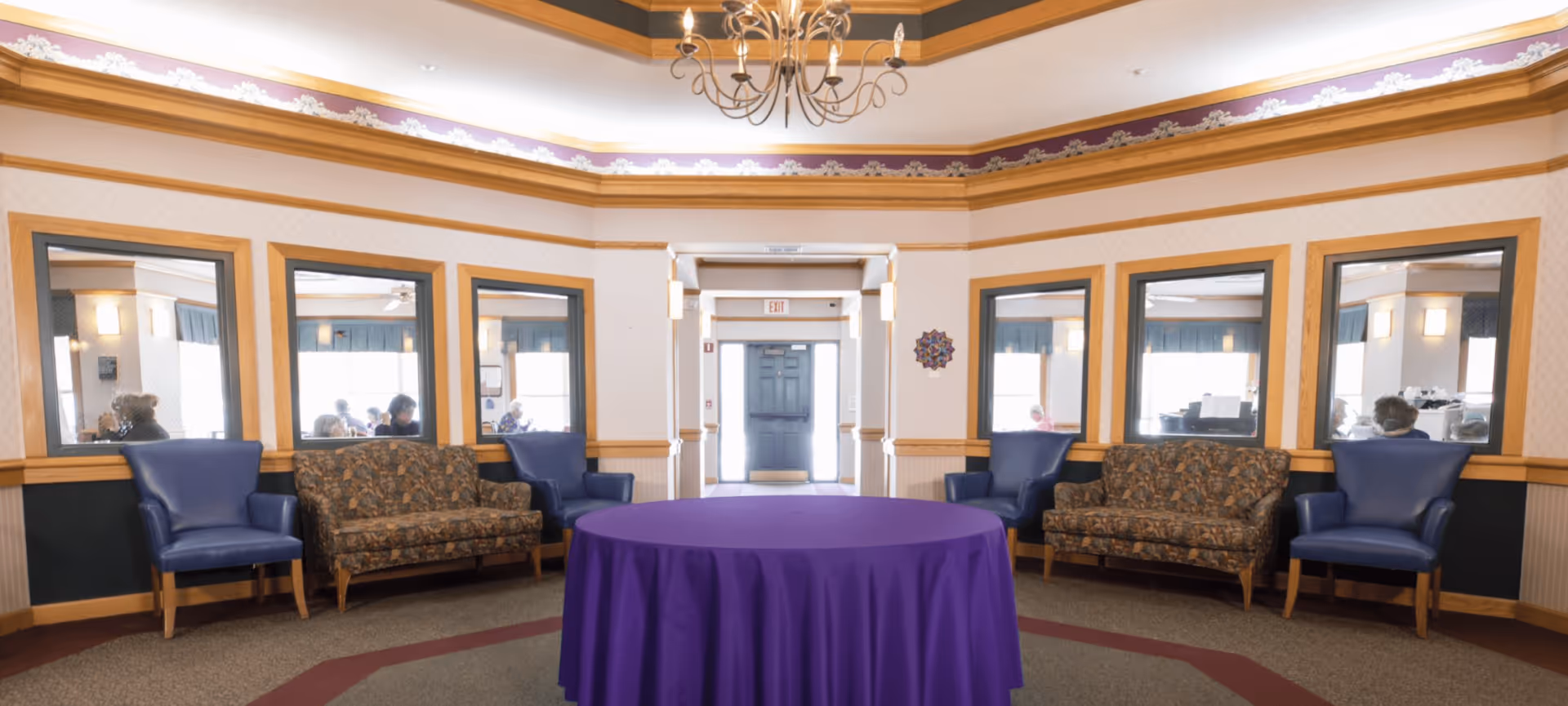 A communal seating area with armchairs and patterned sofas arranged around a round table draped in a purple tablecloth under a chandelier.