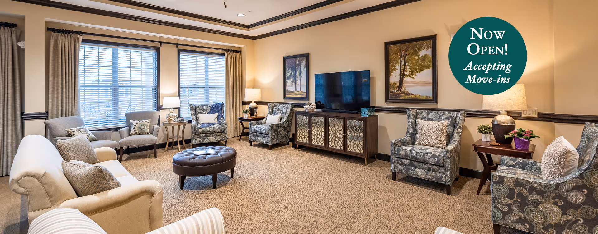 Bright senior living room with sofas, armchairs, ottoman, TV console and framed art, plus a promotional 'Now Open! Accepting Move-ins' badge.