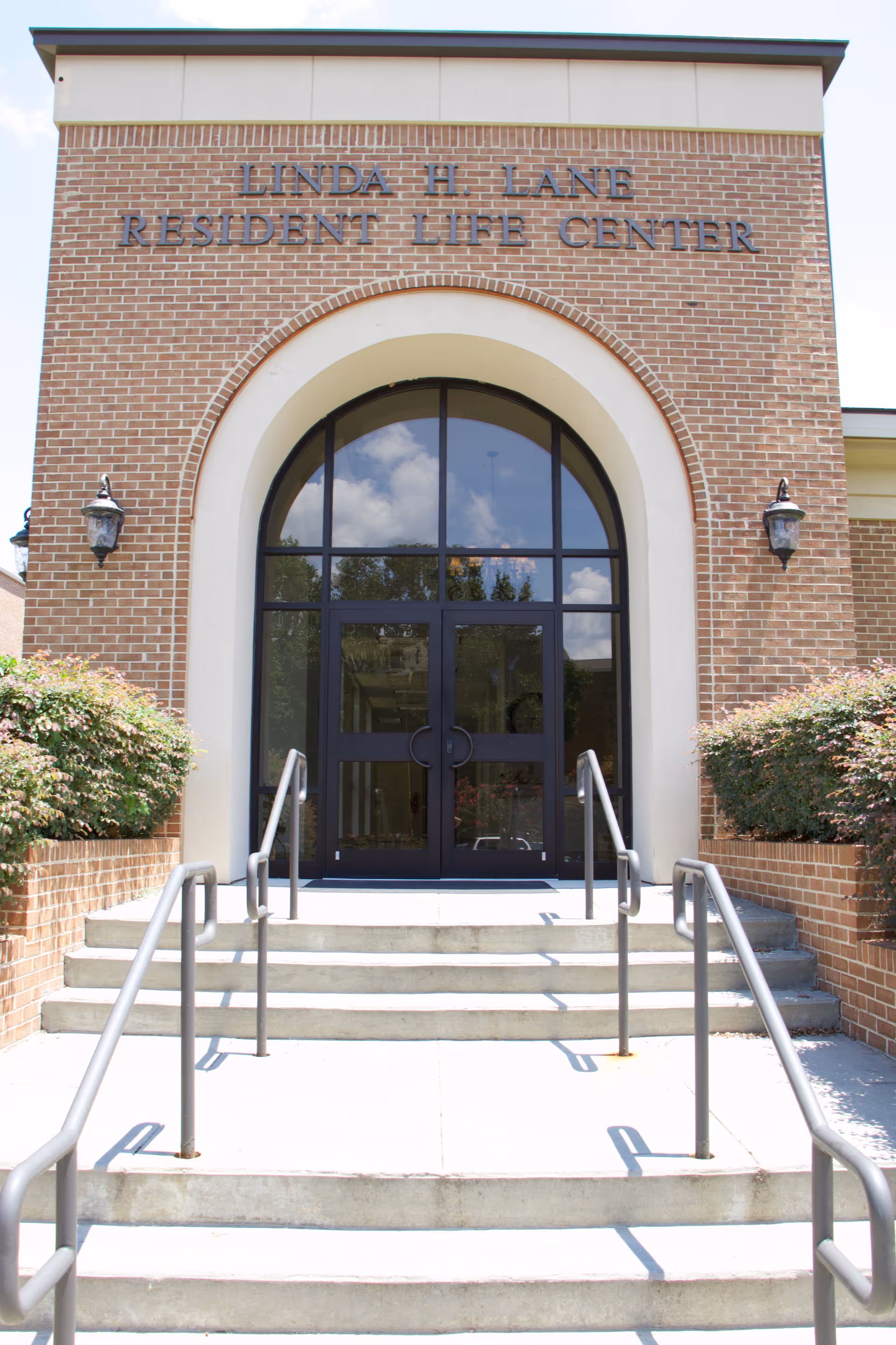 Entrance to the Linda H. Lane Resident Life Center, featuring a brick facade with an arched doorway and glass double doors. There are steps leading up to the entrance with metal handrails on both sides, and bushes flank the stairs.