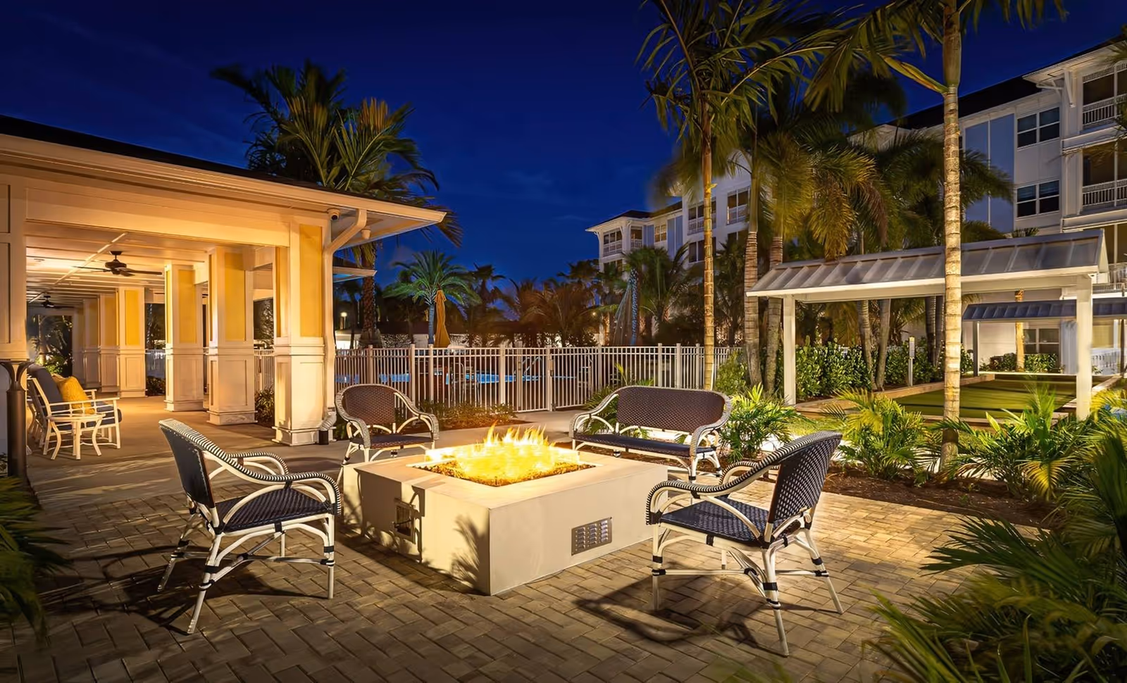 Outdoor evening courtyard with a central square fire pit surrounded by wicker chairs, palm trees, and a covered seating area in front of a multi-story building.