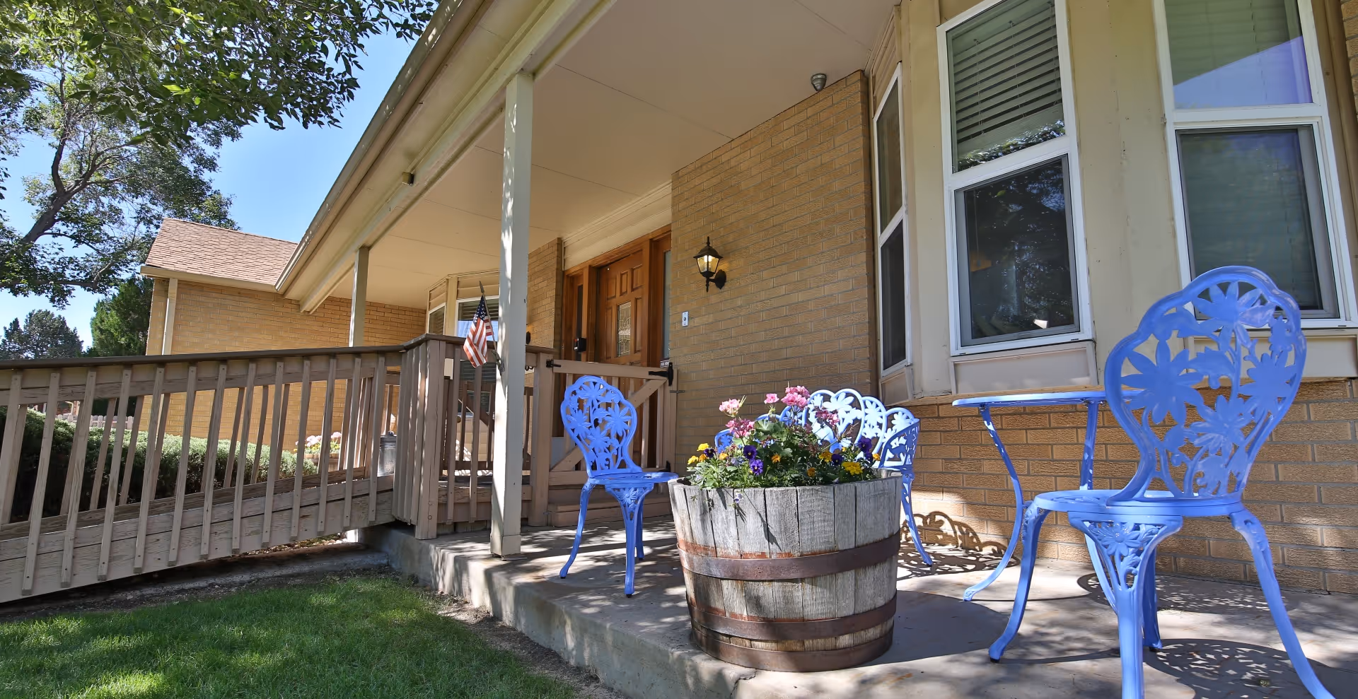 Outdoor patio area at Lighthouse Assisted Living - Elizabeth with blue metal chairs and a small table on a concrete porch. A wooden planter filled with colorful flowers is placed in front of the chairs. The porch has a wooden ramp and steps leading to a door under a covered roof. The building exterior is made of light brown brick with white-framed windows.