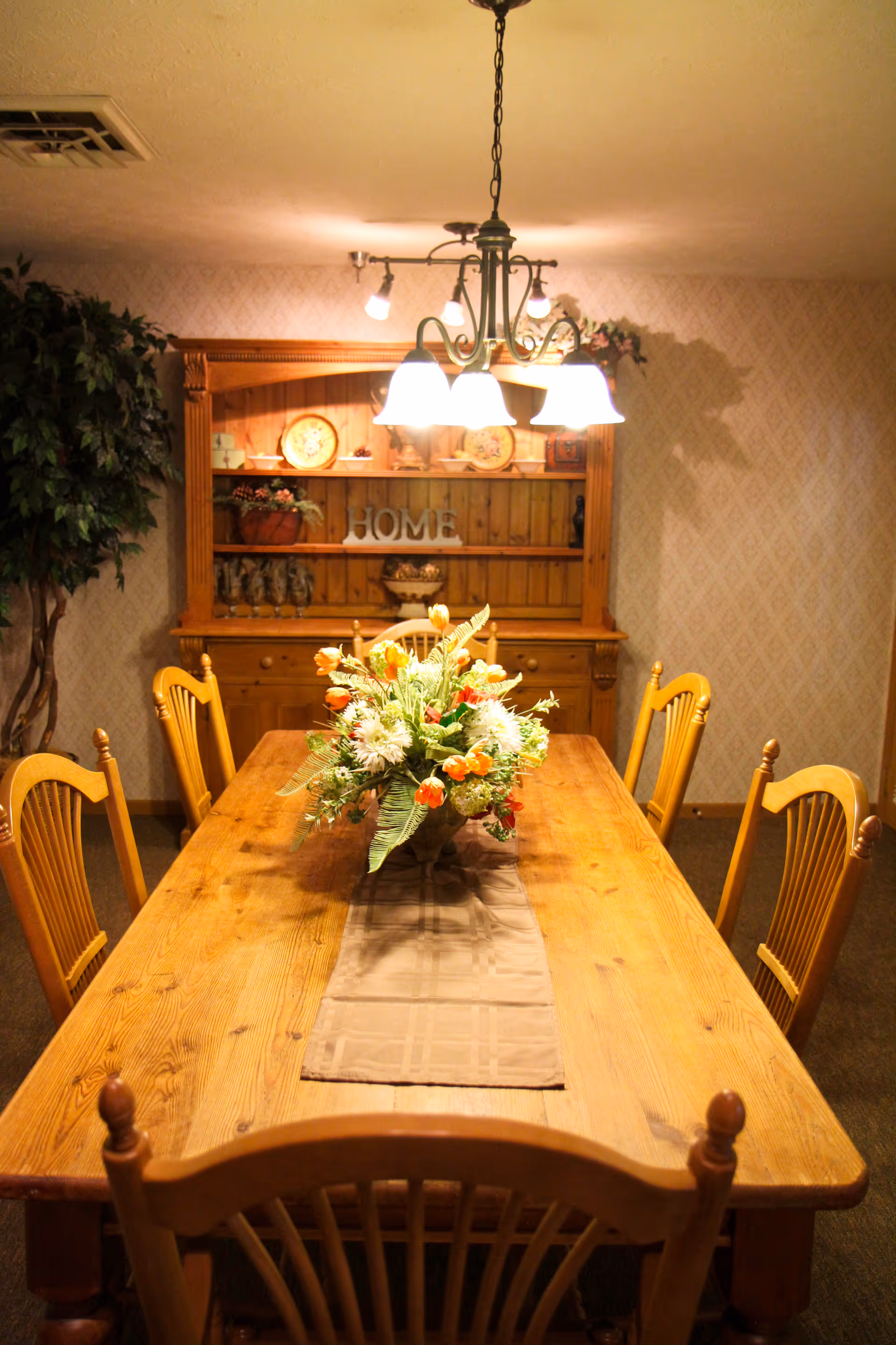 A warmly lit dining room with a wooden dining table and six matching chairs. A floral centerpiece with orange and white flowers sits on a beige table runner. In the background, there is a wooden hutch with decorative plates, plants, and the word 'HOME' displayed. A chandelier with five lights hangs above the table, and a large potted plant is visible to the left.
