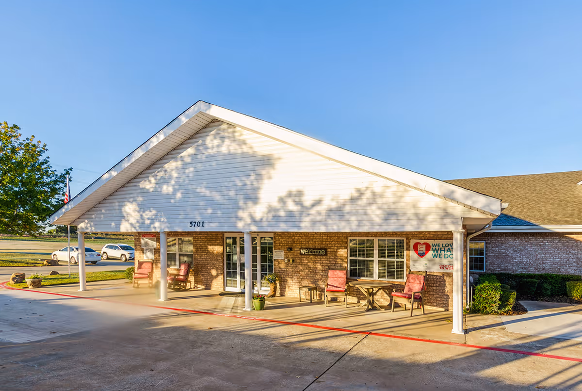 Front entrance of a single-story brick senior living building with a covered portico, outdoor seating, and welcome signage.