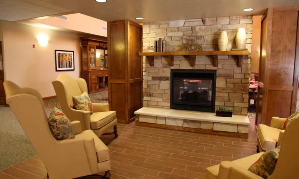 A cozy seating area with four beige armchairs arranged around a stone fireplace with a wooden mantel. The mantel is decorated with vases and books. The room has warm lighting, wooden paneling, and a carpeted area visible in the background with framed artwork on the wall.