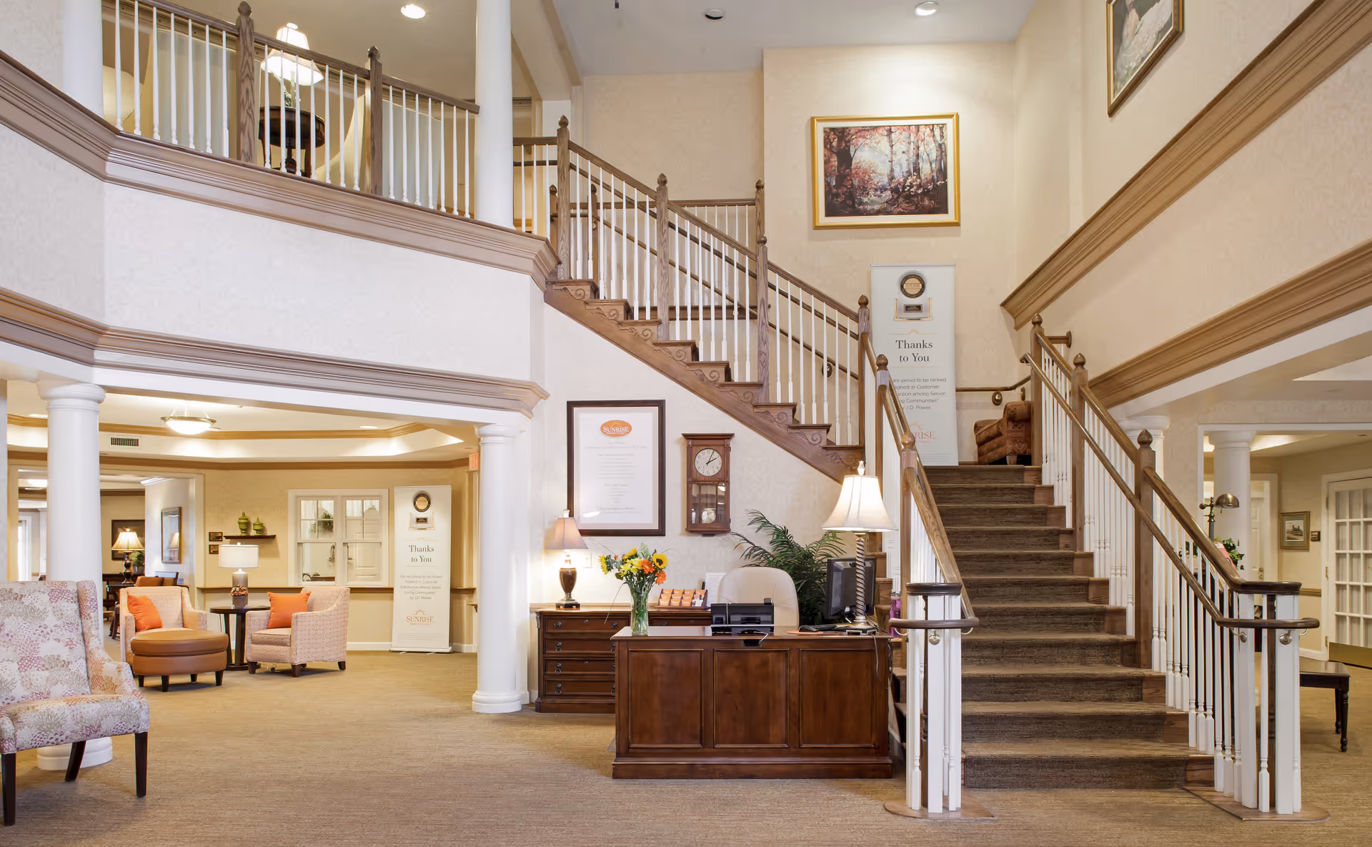 Interior view of a senior living facility lobby with a wooden reception desk, a staircase with wooden handrails and white balusters, comfortable seating area with chairs and ottomans, decorative plants, framed artwork on the walls, and warm lighting.