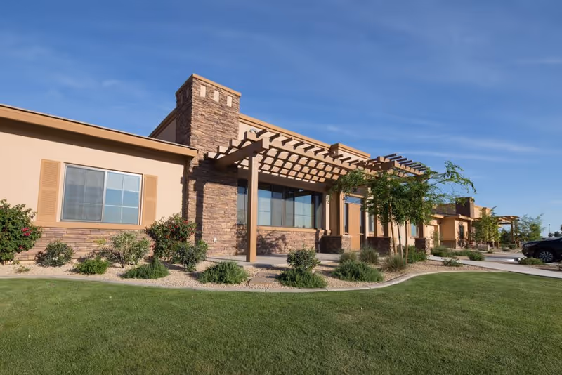 Exterior view of a single-story senior living facility building with beige walls, stone accents, large windows, and pergolas. The building is surrounded by well-maintained green grass, shrubs, and small trees under a clear blue sky.