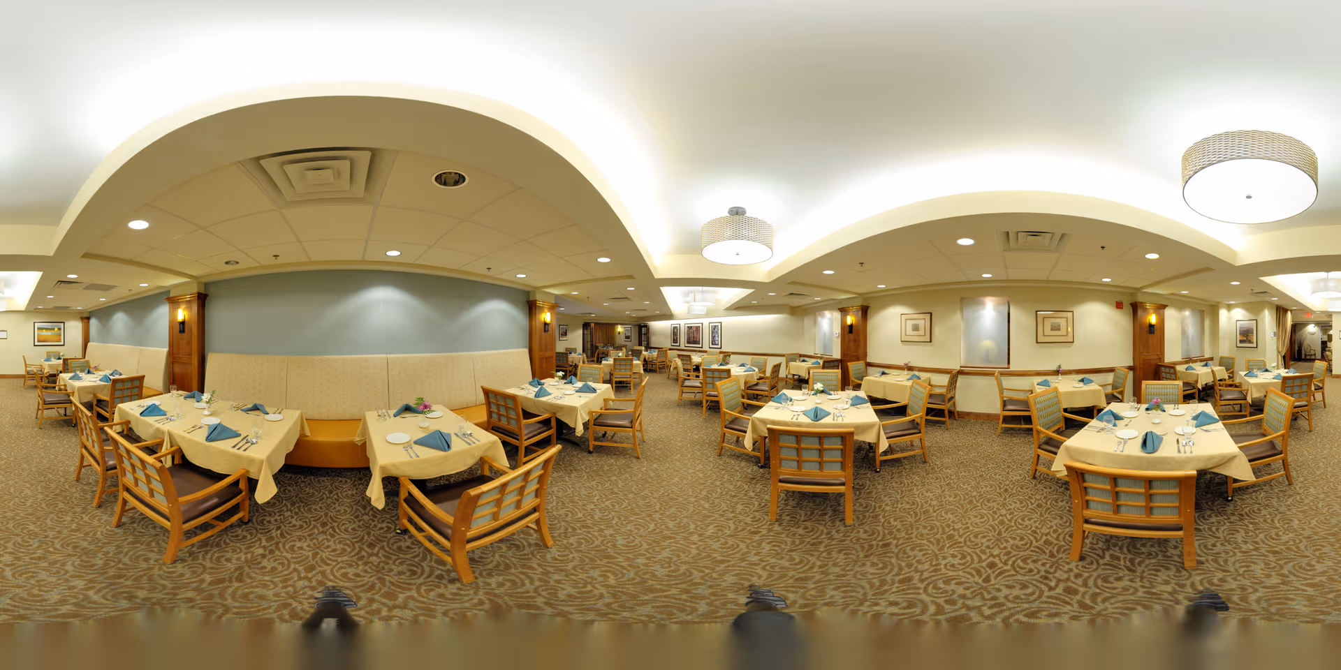 A spacious dining room in The Montebello on Academy with multiple tables covered in beige tablecloths, each set with blue folded napkins, plates, glasses, and silverware. The room features wooden chairs with cushioned seats, patterned carpet, soft lighting from ceiling fixtures, and framed artwork on the walls.