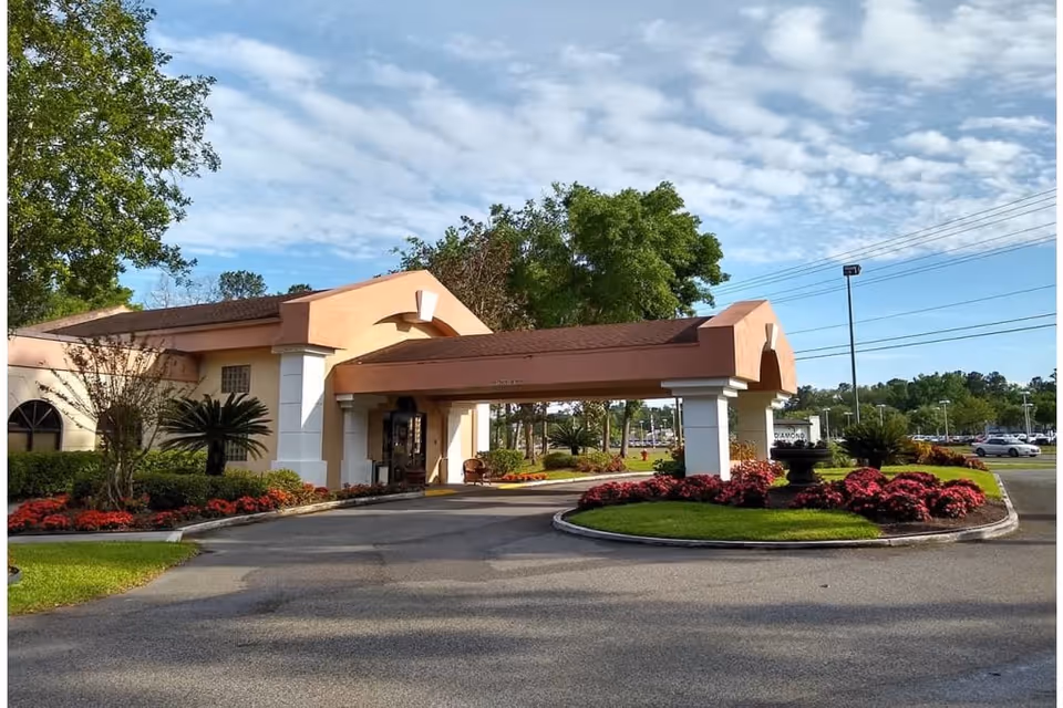 Exterior view of Diamond Assisted Living & Memory Care facility entrance with a covered driveway, landscaped flower beds with red flowers, green grass, and trees under a partly cloudy sky.