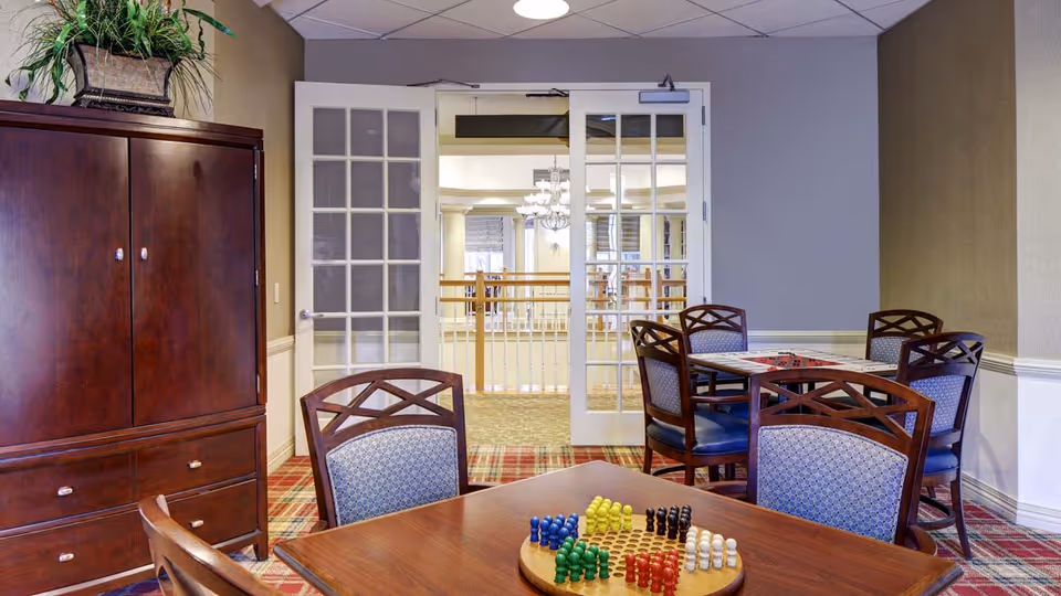 A cozy interior room with wooden furniture including a cabinet and two tables with chairs. One table has a Chinese checkers game set up with colorful pieces. The room has a patterned carpet and beige walls. French doors with glass panes open to a bright area with a chandelier and railing visible in the background.