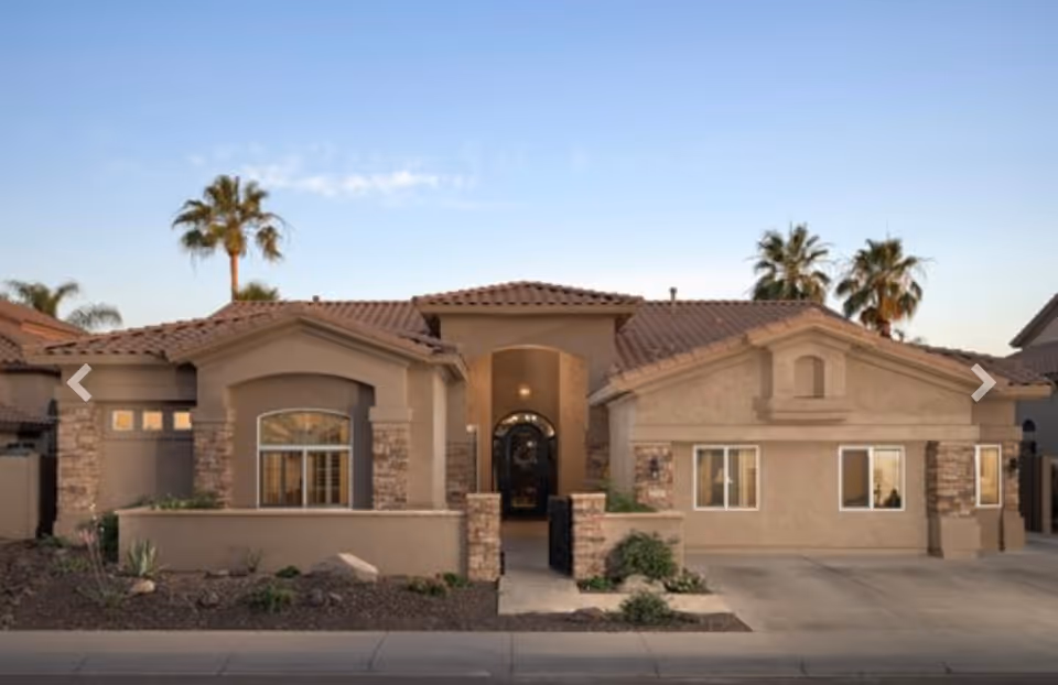 Front exterior view of a single-story residential building with a tiled roof, beige stucco walls, stone accents around windows and entrance, and palm trees in the background under a clear sky.