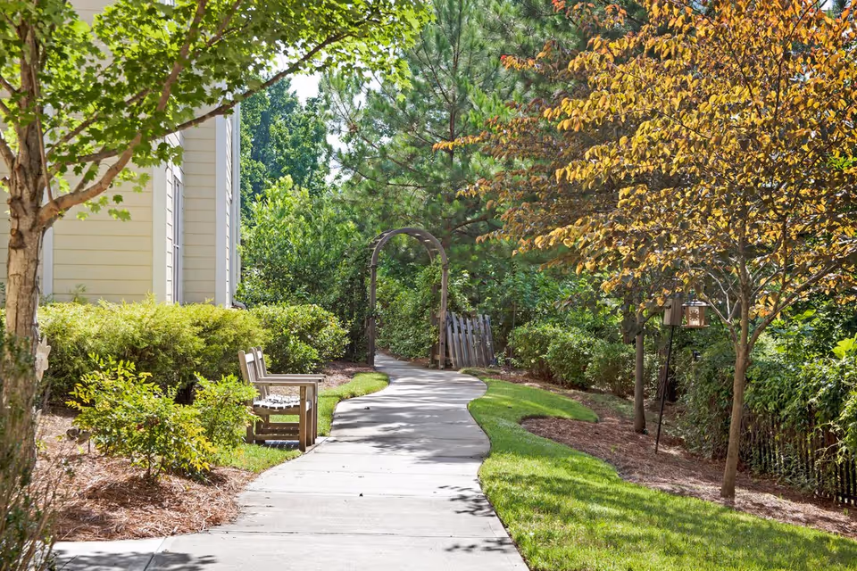 A winding concrete pathway through a garden area with green grass, bushes, and trees. There is a wooden bench on the left side near a building, and a wooden archway further down the path surrounded by greenery.