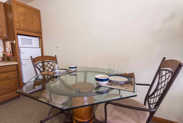 A small dining area with a glass-top table set for two, featuring two chairs with cushioned seats and metal backs. On the table are two bowls, two plates on placemats, and two drinking glasses. In the background, there is a wooden cabinet and a white microwave and refrigerator unit.