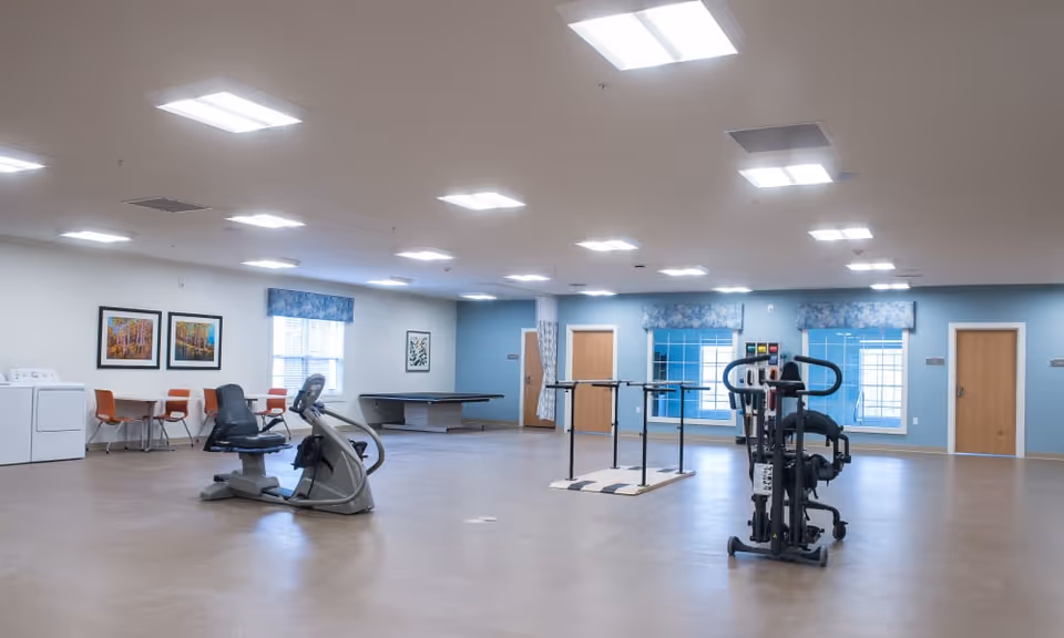 A spacious rehabilitation room with exercise equipment including a stationary bike and parallel bars. The room has light blue walls, several windows with blue floral valances, and bright overhead lighting. There is a small seating area with orange chairs and a table, a laundry area with a washer and dryer, and a pool table in the background.