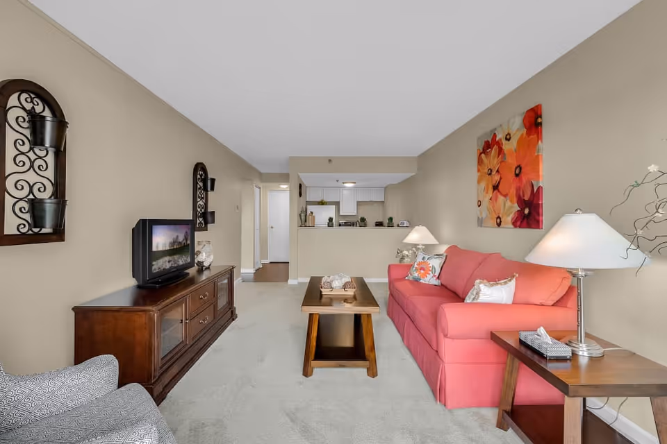 A cozy living room with a coral pink sofa adorned with decorative pillows, a wooden coffee table, and a side table with a lamp and tissue box. Across from the sofa is a wooden TV stand with a television and decorative items. The walls are beige with floral artwork above the sofa and two decorative wall hangings. The room opens into a small kitchen area with white cabinets and a countertop.