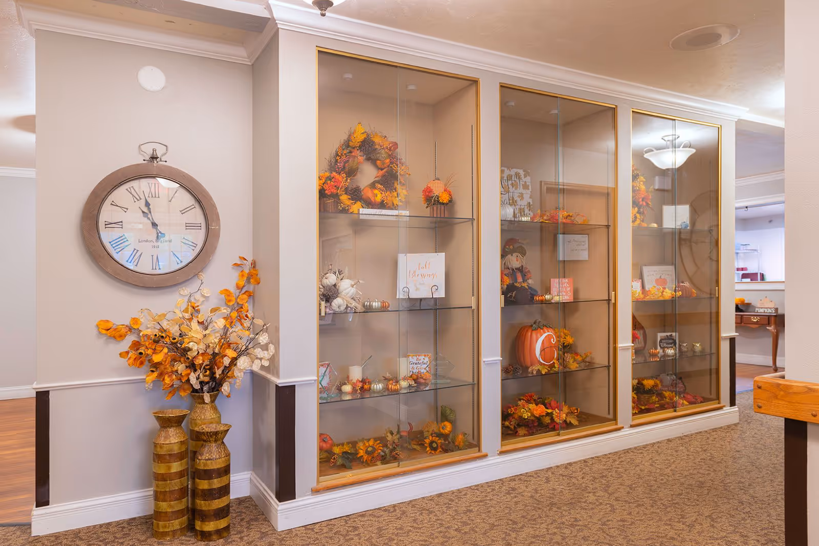 Autumn-themed glass display cabinets and seasonal decorations in a well-lit interior common area with a wall clock and decorative vases.