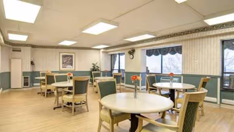 Senior living dining area with round tables, chairs, and flower centerpieces near windows.
