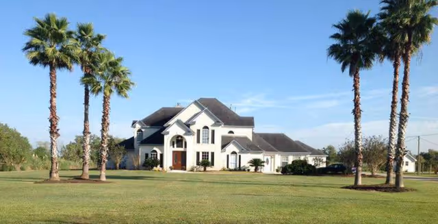 A large two-story white house with a dark roof situated on a spacious green lawn. There are six tall palm trees in the foreground, three on each side of the house. The sky is clear and blue.