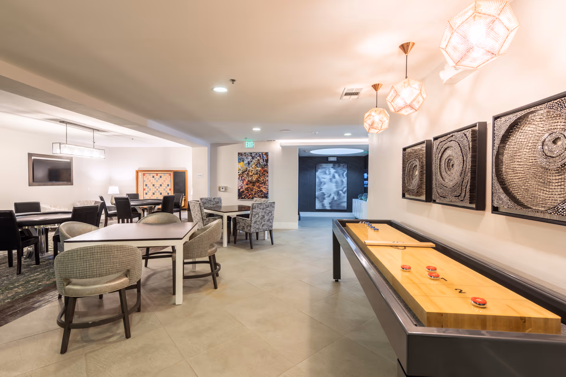 Bright communal interior with multiple tables and chairs and a shuffleboard table beneath pendant lights.