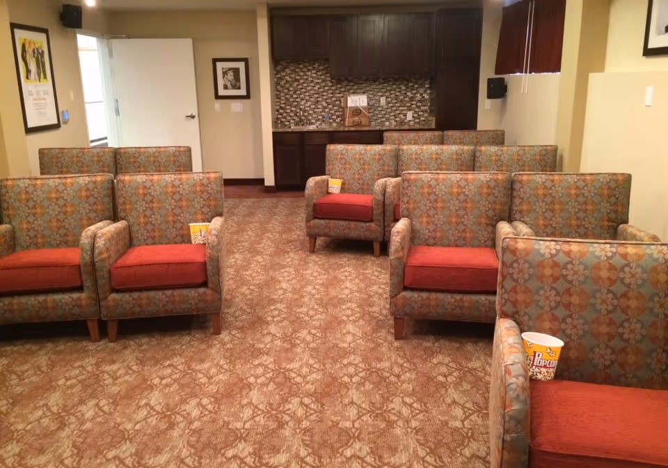 A small theater or media room with patterned armchairs arranged in rows facing forward. Each chair has a red cushion and some have popcorn containers placed on the armrests. The room has a carpeted floor with a geometric pattern, beige walls, a small kitchenette with dark cabinets and a tiled backsplash at the back, and a closed white door on the left side.