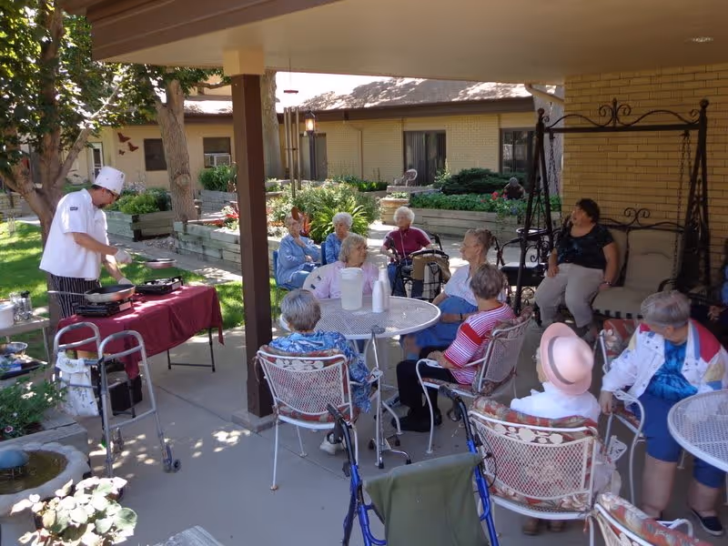 A group of elderly people sitting outdoors under a covered patio at The Courtyard of Loveland Assisted Living. They are seated around metal tables and chairs, some using walkers. A chef wearing a white uniform and hat is cooking on a portable stove at a table covered with a red cloth. The background shows garden beds, trees, and a building with windows.