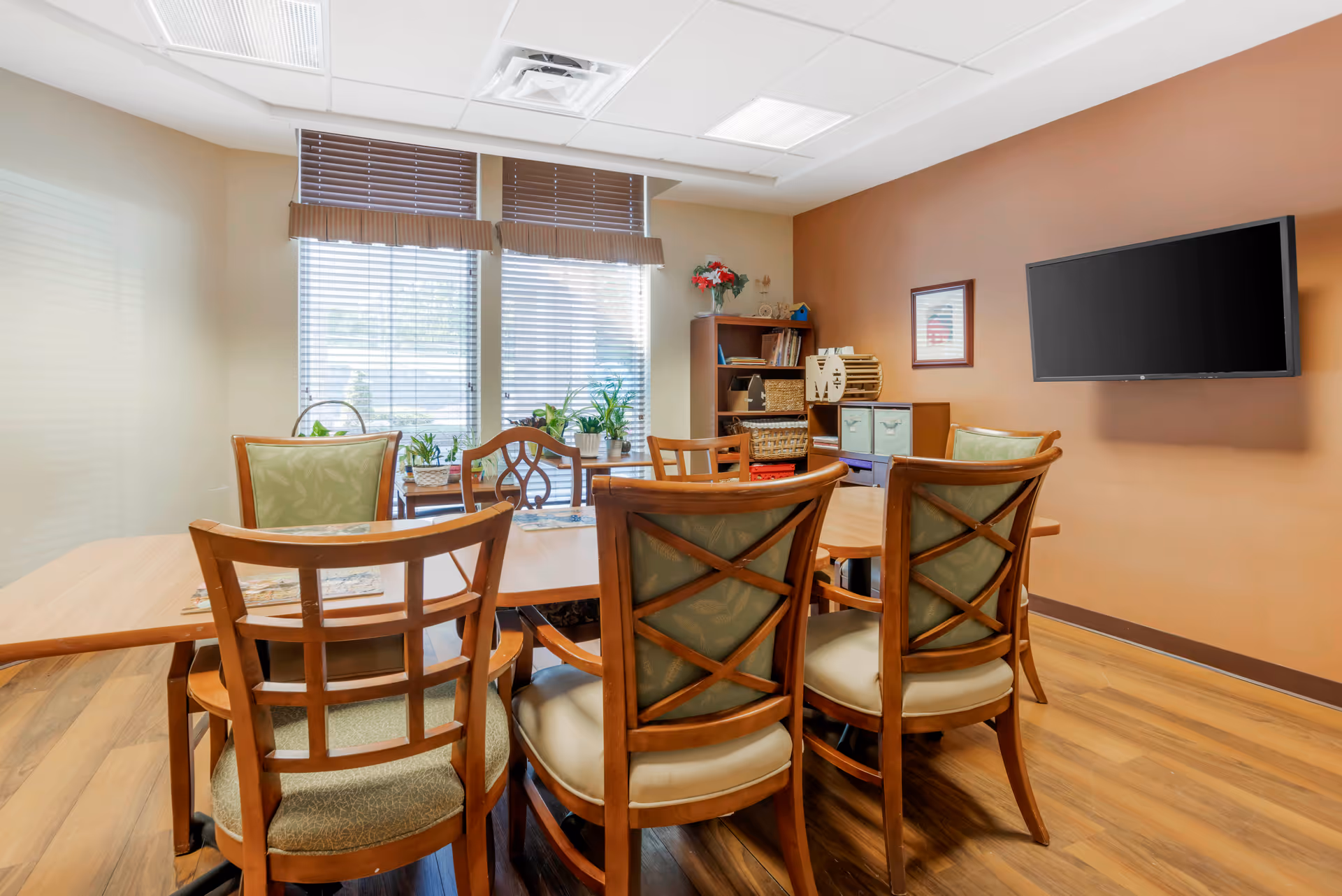 A cozy room with a wooden table surrounded by six chairs with cushioned seats and backs. The room has large windows with blinds allowing natural light to enter. There are several potted plants on a small table near the windows. A bookshelf with baskets and decorative items is against the wall, and a flat-screen TV is mounted on the adjacent wall. The walls are painted in warm tones, and the floor has a wood finish.