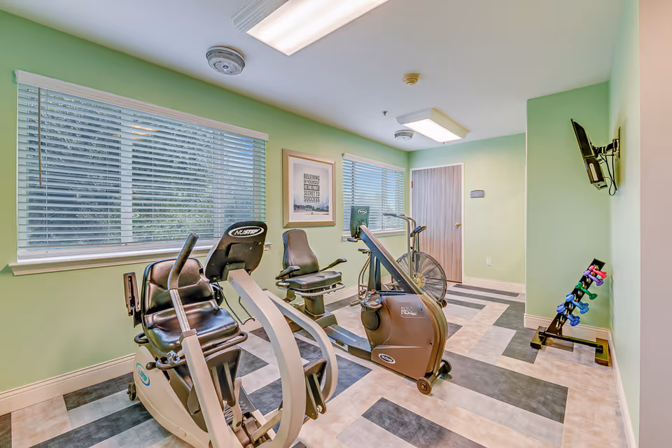 A small fitness room with light green walls and two large windows with blinds. The room contains exercise equipment including a recumbent bike, an air bike, and a rowing machine. There is a rack of colorful dumbbells against the right wall and a wall-mounted TV. A framed motivational poster is hung between the windows.