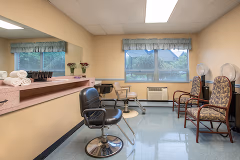 Interior view of a room with salon chairs and hair drying stations. There are two salon chairs in the foreground, a large mirror on the left wall with towels and hair products on the counter, and two hair drying chairs with hoods on the right side near a window with blue valances. The floor is shiny and light blue, and the walls are painted beige.