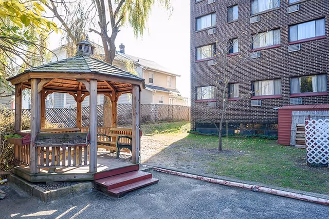 Outdoor area featuring a wooden gazebo with a shingled roof and built-in benches, situated on a paved surface next to a grassy patch. In the background, there is a multi-story brick building with several windows and a neighboring house with light-colored siding. A small tree is planted near the building.