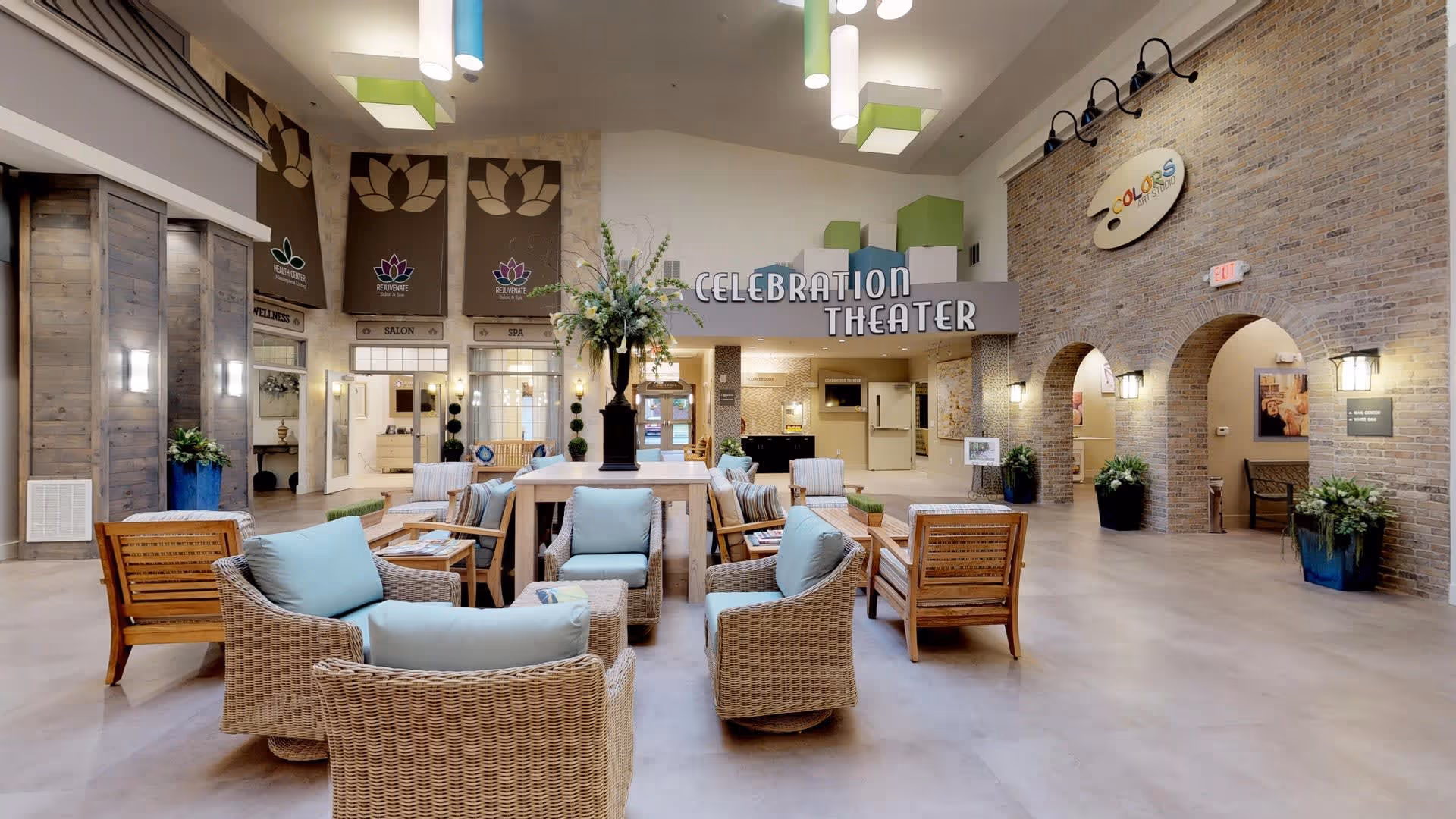 A spacious and well-lit common area in a senior living facility with wicker chairs and wooden armchairs arranged around a central table with a large floral arrangement. The walls feature signs for a salon, spa, and a theater named 'Celebration Theater'. The area has high ceilings with modern hanging lights, brick archways, and decorative plants.