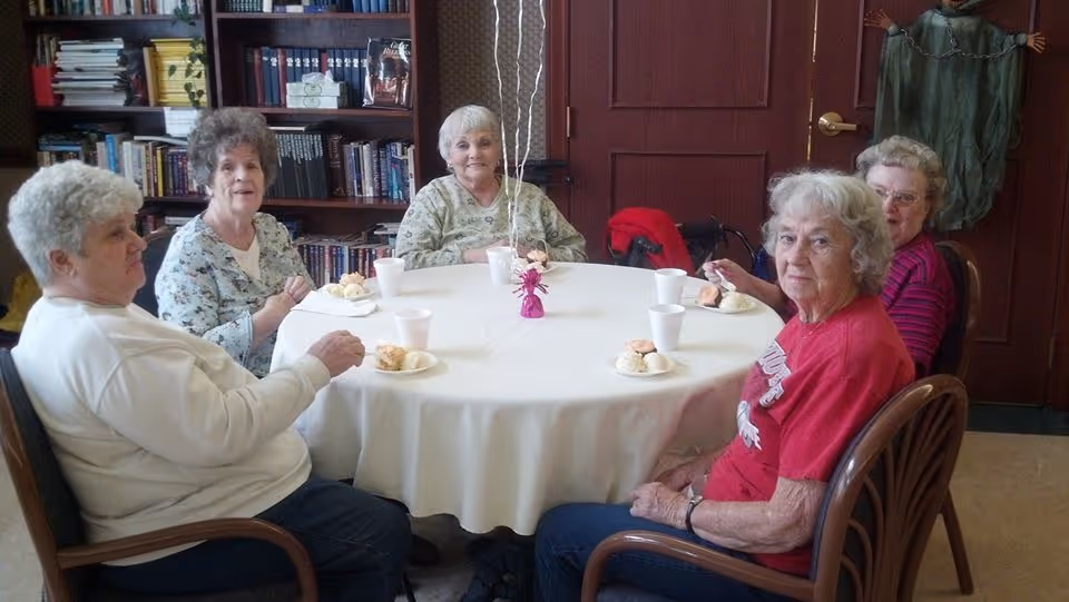 Five elderly women sitting around a round table covered with a white tablecloth, each with a plate of food and a white cup. The background shows bookshelves filled with books and a closed wooden door with a decorative hanging.