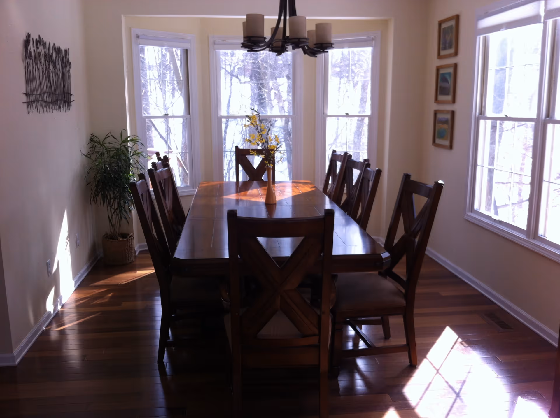 A dining room with a long wooden table surrounded by eight matching wooden chairs. A vase with yellow flowers is placed in the center of the table. The room has large windows on two walls letting in natural light, wooden flooring, a potted plant in the corner, and framed pictures on one wall. A chandelier with multiple lights hangs above the table.